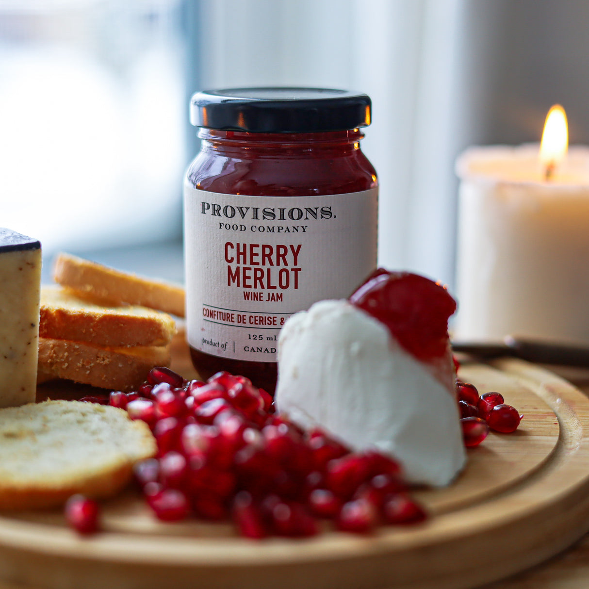 A jar of Provisions Food Company Cherry Merlot wine jam sits on a wooden board with bread, a dollop of cream cheese, pomegranate seeds, and a lit candle in the background.