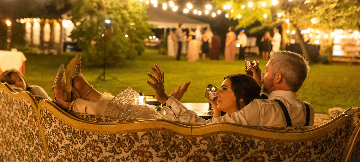 People sitting on a decorative couch at an outdoor event with string lights and greenery.