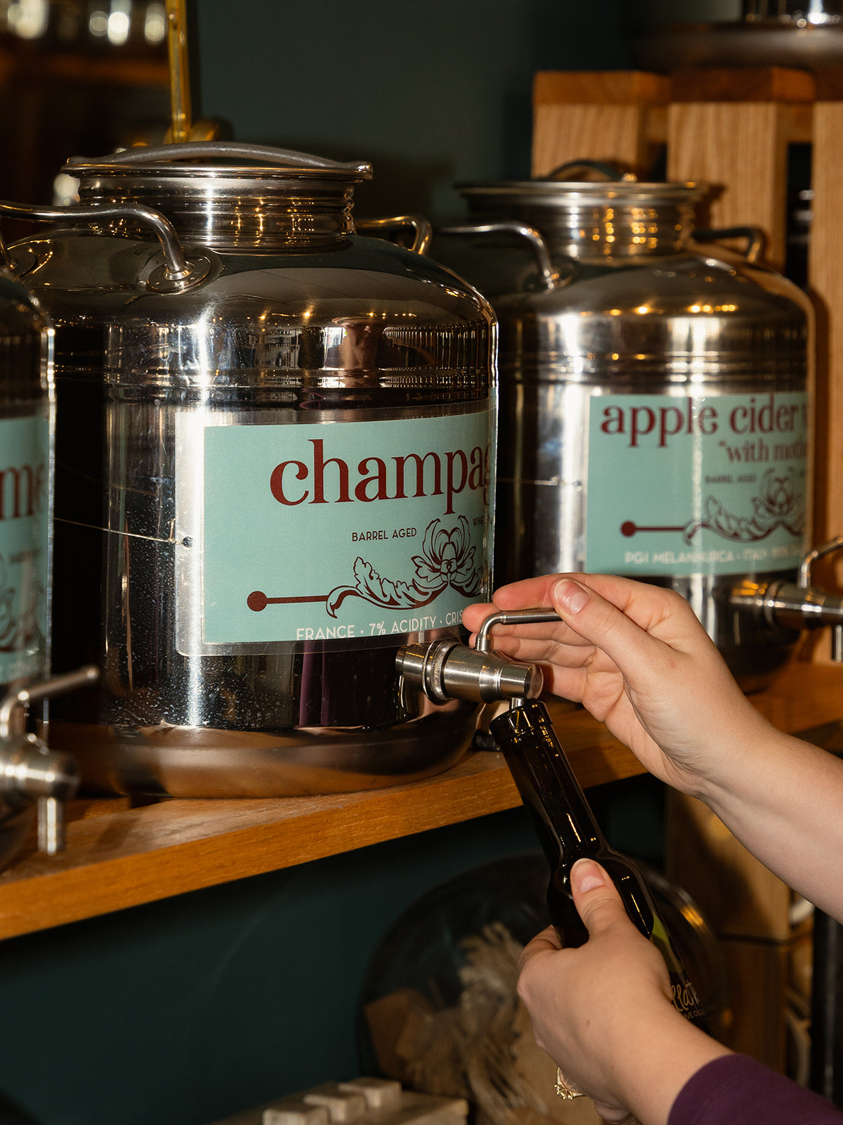 A staff member stands behind a tasting counter in a specialty olive oil and balsamic shop, speaking and gesturing toward shelves lined with stainless steel metal tanks (fusti) labelled with flavours like teriyaki, blackberry, jalapeño lime, garlic cilantro, and black truffle. The video includes close-up shots of the fusti and a stream of dark balsamic being poured into a glass bottle.